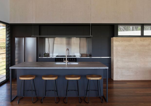 Contemporary kitchen with a long dark island and four timber-seated bar stools, matte dark cabinetry, stainless steel splashback, integrated appliances, and a broad stone-look benchtop. Timber flooring, pale upper cabinetry, and horizontal windows create a refined, warm-toned interior with strong contrast.