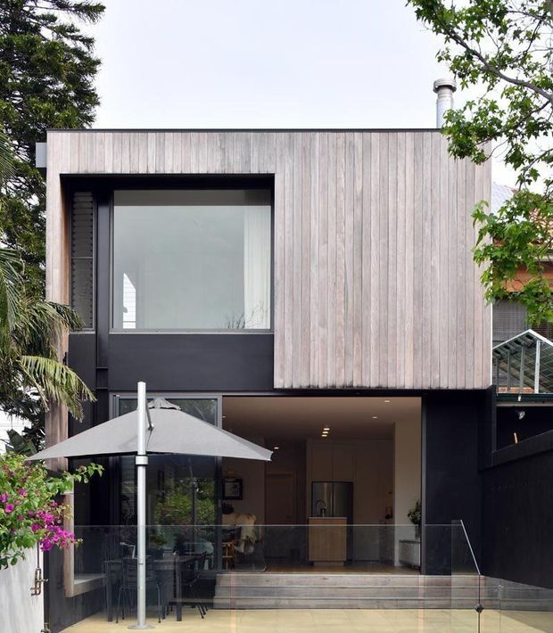 Exterior view of a contemporary two-storey home with a flat roof and a strong geometric facade. The upper level is clad in vertically arranged weathered timber, contrasted with dark painted wall surfaces and large windows. A recessed ground-floor entertaining area opens onto a paved terrace with glass balustrading, outdoor furniture and an umbrella; an interior kitchen or living area is visible through the opening.