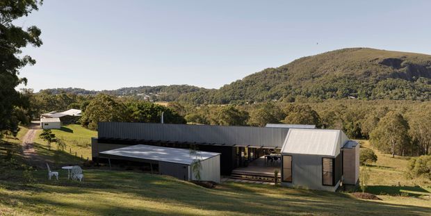 A low-slung contemporary rural residence composed of several linked rectangular volumes, set into a grassy hillside with extensive surrounding bushland and wooded hills. The building has dark corrugated metal wall cladding, pale metal pitched roofs, broad glazed openings, and a covered outdoor terrace or carport-like structure. A smaller outbuilding and outdoor seating are visible to the left.
