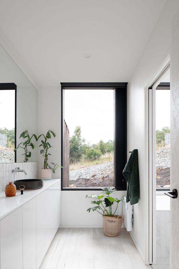 Bright, compact contemporary bathroom with white walls, white vertical wall tiling, pale timber-look flooring, and a wall-mounted white vanity with a dark vessel basin and brushed metal tapware. A large black-framed window provides natural light and overlooks a landscaped construction site, while a black-framed glass door, green towel, and potted plants add contrast and warmth.
