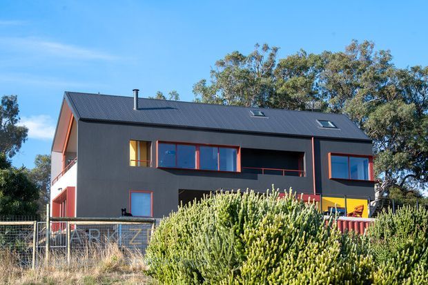 Exterior view of a contemporary multi-storey house with dark charcoal cladding, a pitched metal roof, and bold orange-red window and door frames. The home sits in a landscaped suburban setting with mature trees and dense foreground planting.