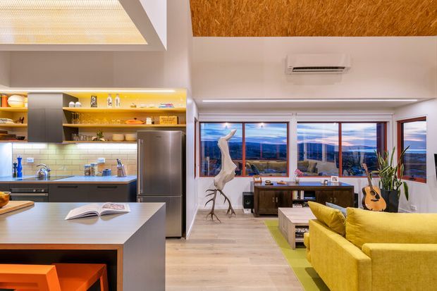 Open-plan contemporary kitchen and living area with a large island, stainless-steel appliances, warm timber open shelving, pale tiled splashback, and light timber flooring. The living zone includes a bright yellow sofa, timber coffee table, desk or media console, indoor plants, and a guitar. Large windows provide expansive views, while a textured timber ceiling panel and recessed lighting add warmth.