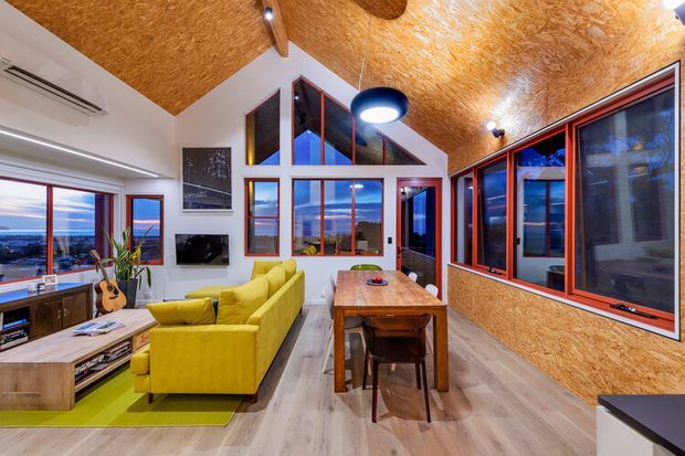 Open-plan living and dining area with a dramatic pitched ceiling clad in warm timber-toned OSB panels, extensive red-framed glazing, and views of the surrounding landscape. The room contains a bright yellow-green sofa, timber media console, dining table and chairs, and pendant lighting. The angular ceiling and high-level glazing create a spacious, contemporary cabin-like interior.