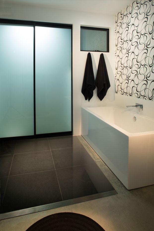 Contemporary bathroom featuring a freestanding white bathtub with integrated chrome wall-mounted tapware, dark large-format floor tiles, a frosted-glass shower enclosure with dark framing, black towels, and a bold black-and-white looping patterned feature wall. Soft under-bath lighting adds a warm glow.
