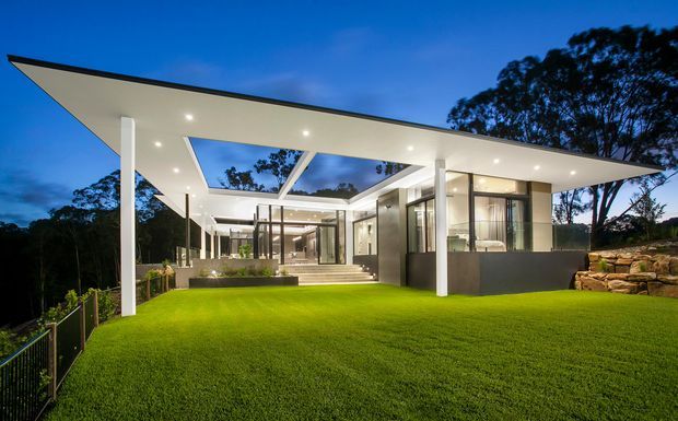 Modern single-storey residence photographed at dusk, featuring a dramatic thin flat roof with broad cantilevered eaves, white structural columns, extensive floor-to-ceiling glazing, and a central open skylit courtyard or roof void. The house opens onto a manicured lawn and landscaped rocky retaining feature, with dark trees surrounding the site.