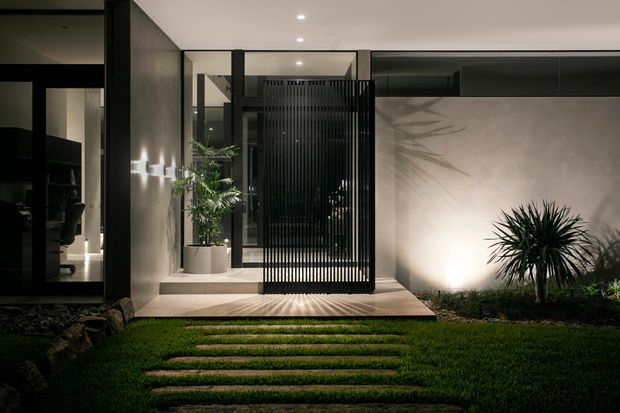Night-time view of a contemporary home entrance opening onto a landscaped garden. The facade combines dark-framed glazing, a pale rendered wall, a recessed entry, and a tall black vertical-slatted screen gate. Stepping stones lead across the lawn to the raised entry platform, with subtle architectural lighting illuminating the walls and planting.
