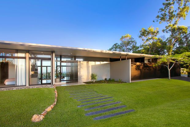 Single-storey contemporary residence with a very low-pitched flat roof, extensive floor-to-ceiling glazing, pale rendered walls, and a covered entrance or carport-like volume. A manicured lawn and irregular dark stepping stones lead toward the glazed living spaces, with mature trees and planting surrounding the building.