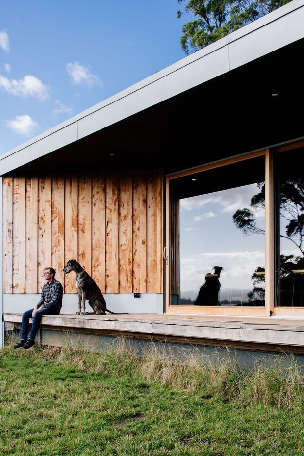 Exterior view of a low-slung contemporary house set in a grassy landscape. The facade combines vertical natural timber cladding with a broad white metal fascia and deep dark soffit. Large timber-framed glazing opens onto a raised timber deck, with a person and dog seated outside. Reflections show trees and distant landscape.
