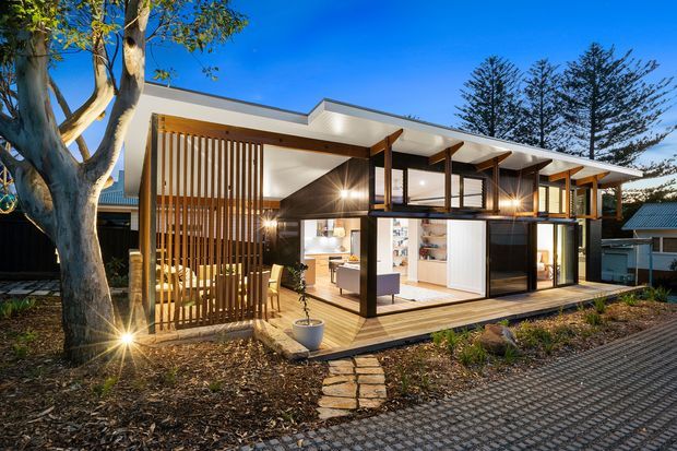 Exterior view of a contemporary single-storey home with extensive glazed openings, dark structural framing, timber decking, vertical timber screening, and a broad low-pitched roof. Warm interior lighting reveals open-plan living areas and fixed cabinetry inside. The house sits in a landscaped suburban setting with a paved approach and mature trees.