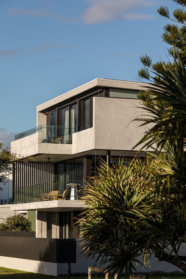 Exterior photograph of a contemporary multi-storey residential building with a flat roof, strongly cantilevered and box-like upper volumes, pale rendered walls, dark vertical screening, extensive floor-to-ceiling glazing, and glass-balustraded balconies. Tropical and subtropical planting partially frames the facade.