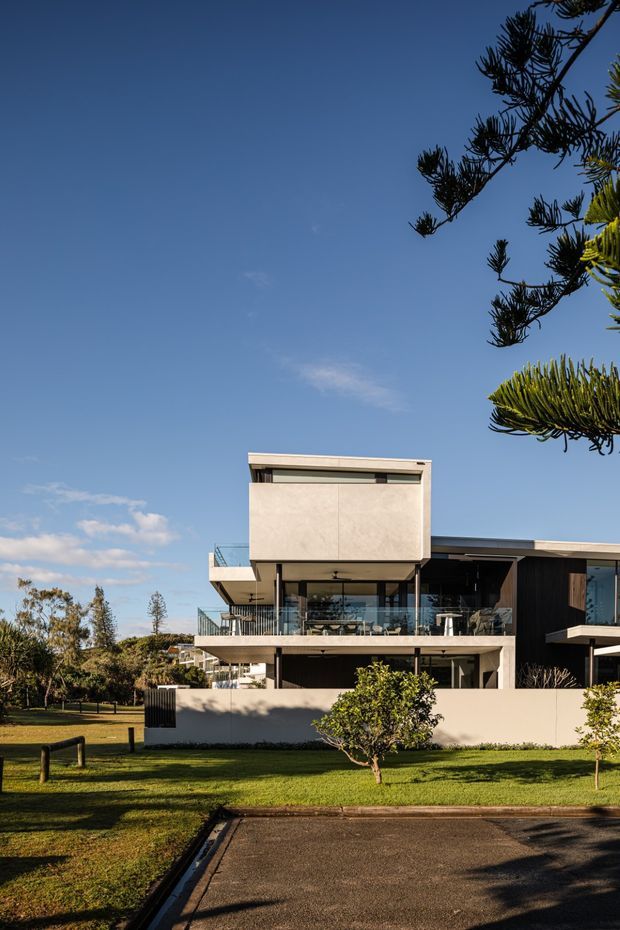 Exterior photograph of a contemporary multi-level residence set beside a landscaped lawn and paved driveway. The building has strong horizontal lines, projecting upper volumes, broad glazing, open balconies with glass balustrades, a flat roof, pale rendered or panelled walls, and dark timber-toned cladding. Mature coastal-looking trees frame the view.