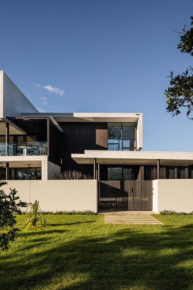 Front exterior view of a contemporary multi-level residence with strong horizontal concrete roof slabs, dark vertical timber or metal screening, expansive glazing, a covered entry canopy, and a slatted pedestrian gate. The home is set behind a pale rendered boundary wall with a neatly landscaped lawn and stepping-stone path.