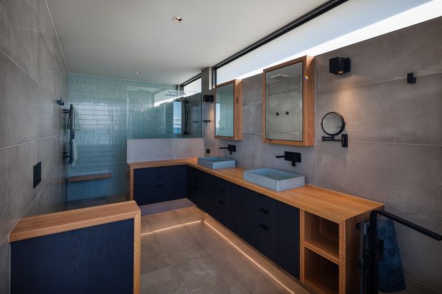 Contemporary double-vanity bathroom with a long timber benchtop, dark built-in cabinetry, two vessel basins, wall-mounted black tapware, framed mirrors, large grey wall and floor tiles, and a glazed shower area at the rear. A built-in timber seat and concealed under-cabinet lighting add warmth and functionality. The room has a sleek, refined, slightly moody atmosphere with strong linear detailing and extensive glazing.