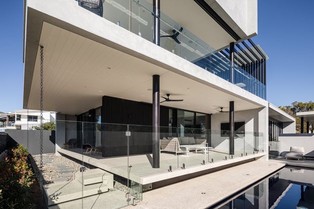 Exterior view of a contemporary multi-level residence with broad cantilevered balconies, white rendered walls, extensive glass balustrades and glazing, dark vertical screening, slender dark columns, and a covered outdoor living terrace beside a swimming pool. The architecture has clean rectilinear lines and strong indoor-outdoor connections.