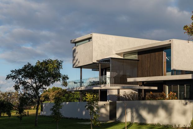 A contemporary multi-level residence viewed from the landscaped front yard. The home features crisp rendered geometric volumes, broad horizontal rooflines, recessed glazing, dark vertical timber cladding, slender metal privacy screens, and a glass balcony. A low rendered boundary wall and mature planting frame the facade under a dramatic cloudy sky.