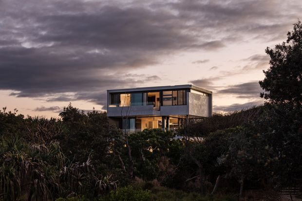 A modern two-storey rectangular house set within dense coastal-looking vegetation, photographed at dusk beneath dramatic cloud cover. The building features a flat roof, extensive dark-framed glazing, a projecting upper level, and warm interior lighting visible through the windows.