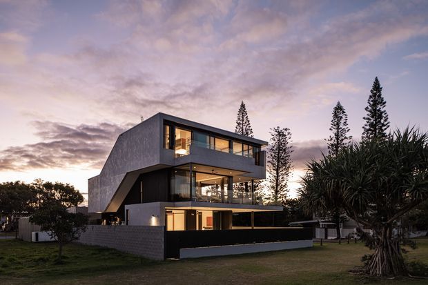 A contemporary multi-storey residence photographed at dusk, featuring a strong geometric form with projecting upper-level volumes, dark cladding, extensive floor-to-ceiling glazing, and warm interior lighting. The house sits within a landscaped suburban setting with mature trees and distinctive palm-like planting, enclosed by a low masonry and dark-panel boundary wall.