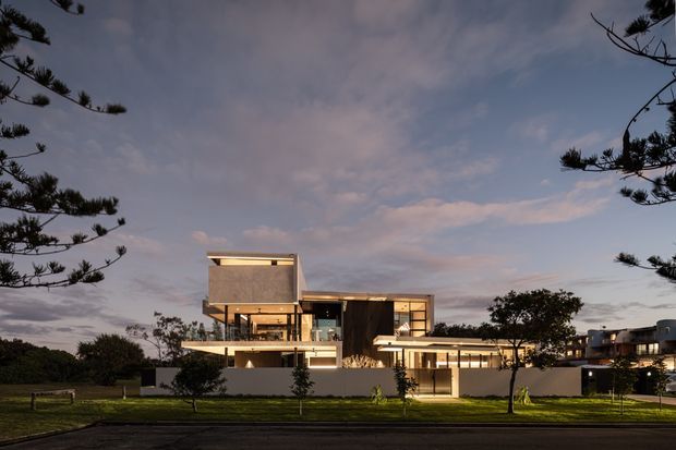 Twilight exterior photograph of a large contemporary multi-level residence, viewed across a landscaped lawn and road. The building has strongly rectilinear volumes, flat roofs, extensive glazing, warm interior lighting, projecting terraces, and a mix of light rendered surfaces and dark vertical cladding. Trees frame the composition, with additional residences visible in the background.