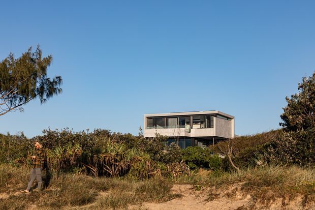A modern two-storey rectangular coastal house set within dense dune vegetation. The building has a flat roof, large horizontal glazing, dark window frames, and pale rendered or concrete walls. A sandy path and native coastal planting occupy the foreground under a clear blue sky.