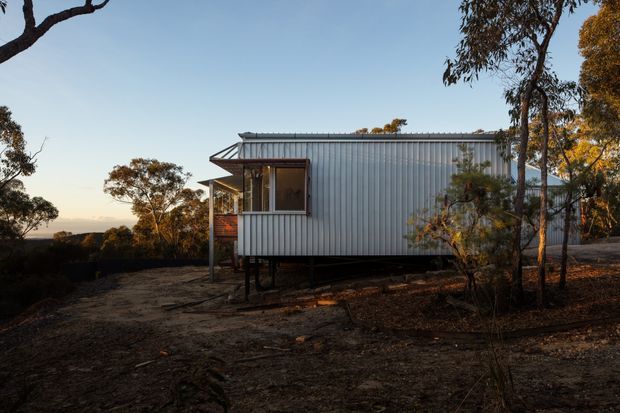 A compact elevated single-storey dwelling set among native bushland, viewed from the side at dusk. The building has a simple rectangular form, corrugated metal wall cladding and roofing, a projecting glazed porch or entry volume, and is supported above the ground on slender posts. The surrounding dry woodland setting and restrained material palette give it a utilitarian, lightweight cabin-like character.