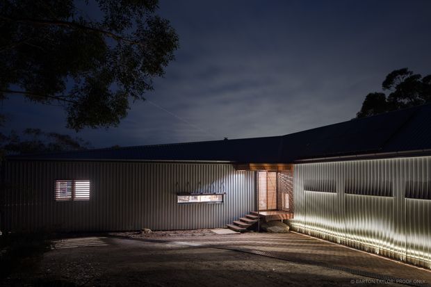 Night-time exterior view of a low, angular building with dark corrugated metal cladding and a dark pitched roof. Linear integrated lighting washes the ribbed facade, highlighting a recessed entrance with timber-toned screening, steps, and a small illuminated window. The building sits beside a paved or gravel forecourt with surrounding trees silhouetted against the sky.