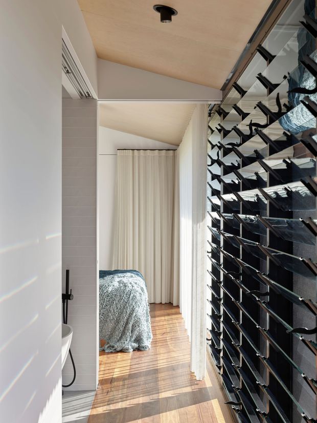 A narrow, sunlit interior passage looking from a bathroom toward a bedroom. The foreground includes a partial white freestanding basin or bathtub with a matte black wall-mounted tap and handheld shower against pale rectangular wall tiles. Beyond, a bed draped in a textured blue-grey throw sits on a warm timber floor, framed by full-height cream curtains. Along the right side is a distinctive black louvred privacy screen with adjustable horizontal blades, and the ceiling has warm timber lining with recessed black lighting.