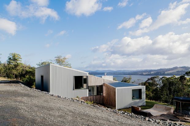 A modern angular house on a sloping site, composed of several connected box-like volumes with corrugated light-grey metal cladding and sections of vertical timber cladding. The building overlooks a broad body of water and distant hills, with large glazed openings and a gravel-covered foreground. Bright daylight and expansive sky emphasize the elevated coastal or lakeside setting.