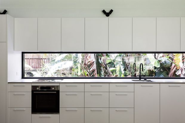 A streamlined contemporary kitchen with extensive white flat-panel cabinetry, integrated upper cupboards, a dark benchtop, stainless-steel sink and tap, and a built-in black oven. A long horizontal window above the benchtop provides strong natural light and overlooks dense tropical greenery. The space has a clean, minimal composition with subtle black track lighting and understated linear handles.