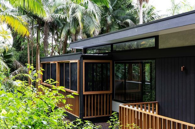 A contemporary dark-clad house nestled among dense tropical vegetation. The single-storey structure has a low-pitched roof, extensive dark-framed glazing, vertical black cladding, and warm timber-framed windows, entry elements, and deck balustrades. The architecture appears integrated into a lush forested setting.
