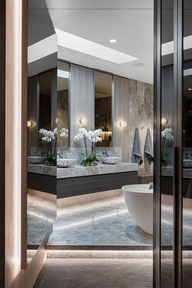 Luxurious contemporary bathroom viewed through a doorway, featuring a double vanity with dark wood cabinetry, a large natural-stone benchtop and matching stone floor, full-height mirrors, vertical timber-toned wall panels, integrated round lighting, orchids, and a freestanding white bathtub. A skylight or recessed ceiling opening provides natural illumination, while illuminated floor-level steps and layered reflections add depth.