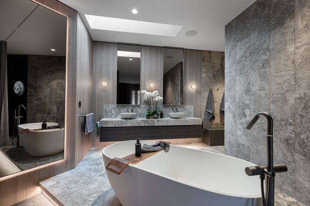 Luxurious contemporary bathroom with a freestanding white bathtub in the foreground, a double vanity with twin basins, large mirrors, warm grey timber-look wall panelling, and full-height grey stone-look wall and floor finishes. A skylight provides natural light, while integrated pendant-style lights and recessed ceiling lighting add a refined atmosphere. The shower and bathtub are visible in the mirror reflection.