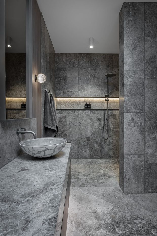 A moody, contemporary bathroom clad extensively in grey veined stone-look tiles. The room features a walk-in shower with dark fixtures, a floating stone vanity with a decorative vessel basin, a large mirror, integrated warm LED shelf lighting, and a towel rail. The palette is predominantly charcoal and mid-grey with subtle warm lighting.