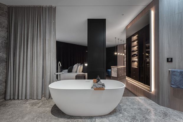 A luxurious contemporary bathroom featuring a freestanding white oval bathtub on a grey stone-look floor, with full-height grey textured wall panels, a large curtain, and a dark timber dressing or wardrobe zone illuminated by integrated lighting. A partial bedroom or lounge area is visible beyond the tub.