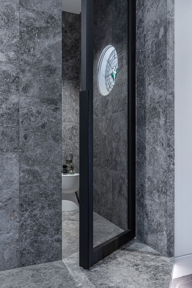 A dramatic, monochromatic bathroom interior clad in large-format grey veined stone tiles. A tall black-framed mirrored door or partition stands partially open, reflecting the tiled space and a white freestanding or pedestal basin. An oval stained-glass window provides a small green and multicoloured accent, while the stone flooring and walls create a luxurious, moody atmosphere.