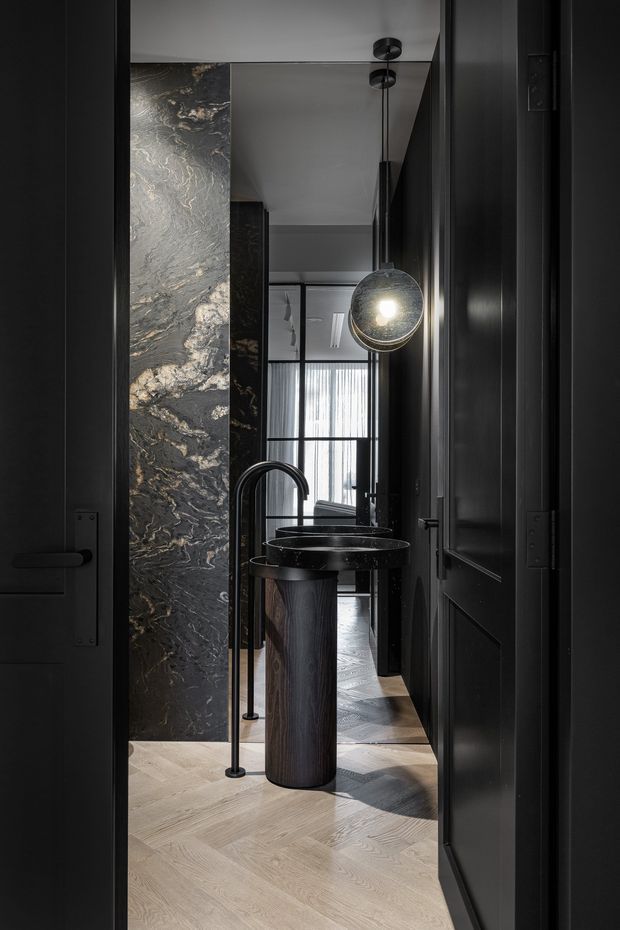 A narrow, dramatically styled bathroom viewed through dark paneled doors. The space features a freestanding dark cylindrical vanity with a round black basin and sculptural black tapware, a dramatic dark veined stone wall, pale herringbone timber flooring, black-framed glazing, and a spherical pendant light. The palette is predominantly black, charcoal, grey, and light timber, creating a refined, moody atmosphere.
