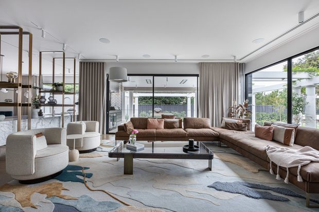 A spacious contemporary living room with extensive floor-to-ceiling glazing, sheer curtains, a large brown sectional sofa, sculptural upholstered armchairs, an open metal-and-wood display shelving unit, and a low rectangular coffee table. The room has a light neutral palette with layered organic-shaped rugs and views to a landscaped exterior courtyard or garden.