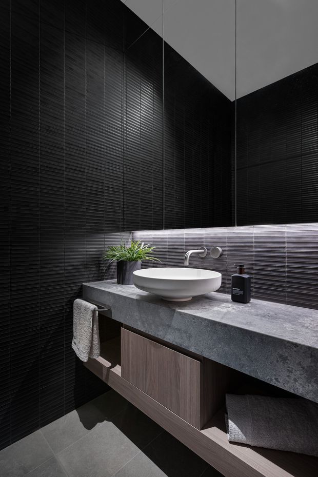A dramatic contemporary bathroom vanity featuring dark horizontally ribbed wall tiles, a floating timber-toned cabinet with open towel storage, a thick grey stone benchtop, a white vessel basin, and wall-mounted chrome tapware. A small potted plant and toiletries sit on the counter, with grey floor tiles completing the moody scheme.