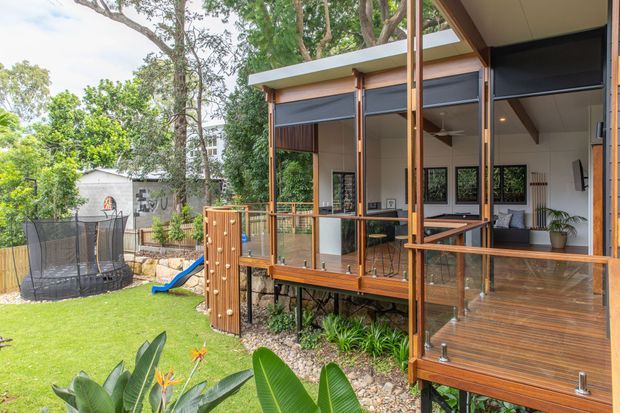 Exterior view of a raised contemporary timber-clad home surrounded by established greenery. The image focuses on a generous timber deck and covered outdoor living area with glass balustrades, exposed timber posts and roof framing, and large glazed openings into a bright interior. A lawn and landscaped garden occupy the foreground, with a small play structure visible to the side.