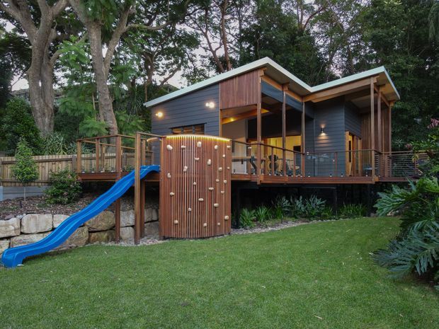 Exterior view of a raised contemporary timber house set within a leafy garden. The dark horizontal cladding, angular skillion-style roof forms, warm timber screening, covered deck and large glazed openings are prominent. A blue children's slide connects to the elevated deck, with stone retaining walls and landscaped planting in the foreground.