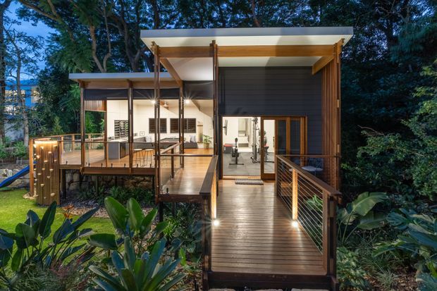 A modern elevated timber home nestled in dense greenery, photographed at dusk. A broad timber walkway and bridge-style deck lead to a glazed entry, with a covered outdoor terrace and dining area extending to the left. The structure features slender dark posts, warm timber soffits and decking, dark horizontal cladding, cable balustrades, and generous openings connecting the interior with the surrounding garden.