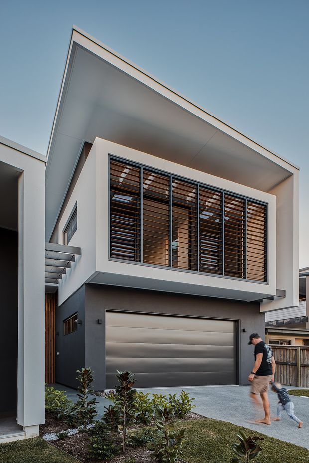 A contemporary two-storey residential facade viewed from the street, featuring crisp white and charcoal rendered volumes, a broad cantilevered upper level, a deep roof overhang, dark-framed windows with horizontal timber-look louvres, and a metallic sectional garage door. A paved driveway, landscaped planting, and neighbouring homes are visible in the foreground and background.