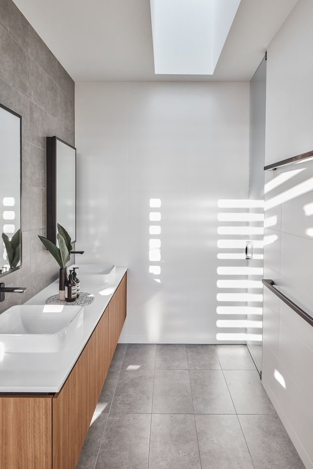 Bright contemporary bathroom with a long timber vanity, white countertop and two white integrated basins. Large-format grey stone-look wall tiles frame a mirror on the left, while white wall tiles and built-in cabinetry line the right side. A large rectangular skylight floods the room with daylight, creating strong horizontal light and shadow patterns.