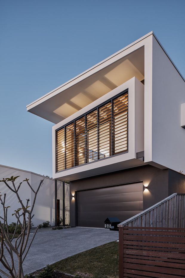 A dusk exterior photograph of a contemporary two-storey residence. The facade features crisp white rendered volumes, a dark grey garage frontage, a projecting flat roof, and a large upper-level window screened with horizontal timber-look louvres. A concrete driveway, timber boundary fence, exterior wall lights, and landscaped frontage are visible.
