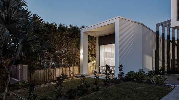 A contemporary residential exterior at dusk, featuring a compact white cubic entry pavilion with a deep recessed porch, tall glazed doorway, dark roller blind, and integrated downlighting. The building is set behind timber fencing and landscaped planting, with a darker glazed volume visible to the right.