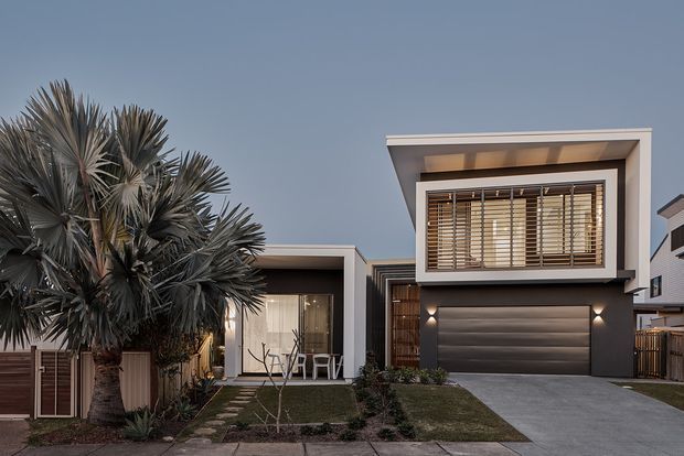 Contemporary multi-level residential facade at dusk, featuring a projecting upper volume with broad timber-look horizontal blinds, a dark garage door, recessed entry, and a low single-storey wing. The home is framed by a mature palm, small landscaped garden beds, lawn, and a concrete driveway.