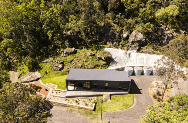 Aerial view of a compact modern single-storey building set within a heavily wooded, rocky landscape. The structure has a long rectangular form, dark metal cladding and a low-pitched roof, with a covered entry terrace, paved paths, landscaped lawn, driveway and several cylindrical outdoor tanks or silos beside it.