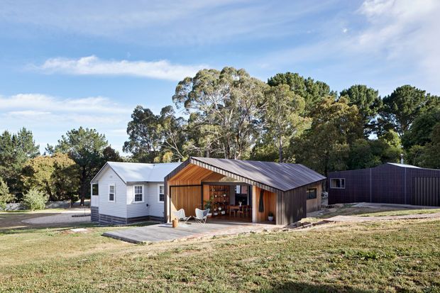 A contemporary rural residential compound set within a landscaped, tree-filled site. The main structure is a compact dark-clad pavilion with a pitched metal roof and a warm timber-lined open living area, positioned beside a small light-coloured weatherboard cottage and a larger dark ancillary building. A concrete path and patio connect the buildings, with expansive lawn and mature trees surrounding the composition.
