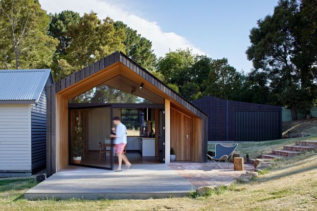 Exterior view of a compact contemporary timber-clad pavilion or extension, featuring a steeply pitched dark metal roof, warm timber soffit and wall lining, broad glazed sliding doors, and a concrete terrace. A small adjoining light weatherboard structure and dark vertical-clad outbuilding sit within a landscaped, wooded site.