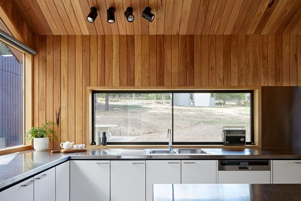 A compact contemporary kitchen with white flat-panel cabinetry, dark benchtops, stainless-steel sink and appliances, and extensive warm timber wall and ceiling lining. A large horizontal window brings in natural light and overlooks an open outdoor setting. Black track spotlights and a small potted plant provide restrained accents.