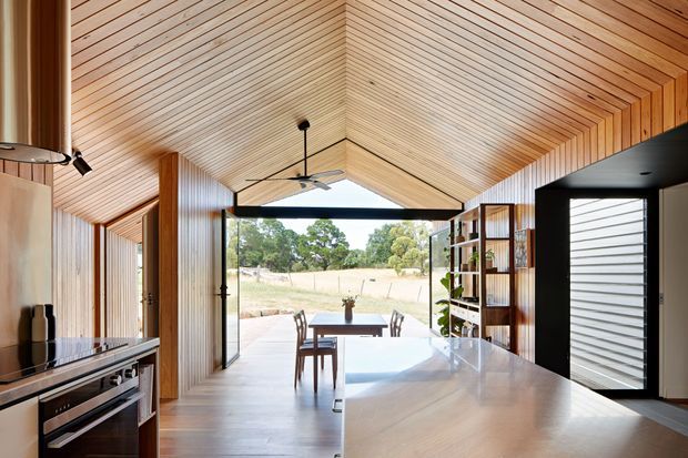 A warm, open-plan kitchen and dining space with extensive vertical timber wall panelling and a vaulted timber-lined ceiling. The kitchen has dark built-in cabinetry and a large light stone island, while full-height glazed doors open to a covered outdoor terrace and rural landscape. A dining table sits centrally beneath a ceiling fan, and a bold black-framed entry or utility zone contrasts with the timber interior.